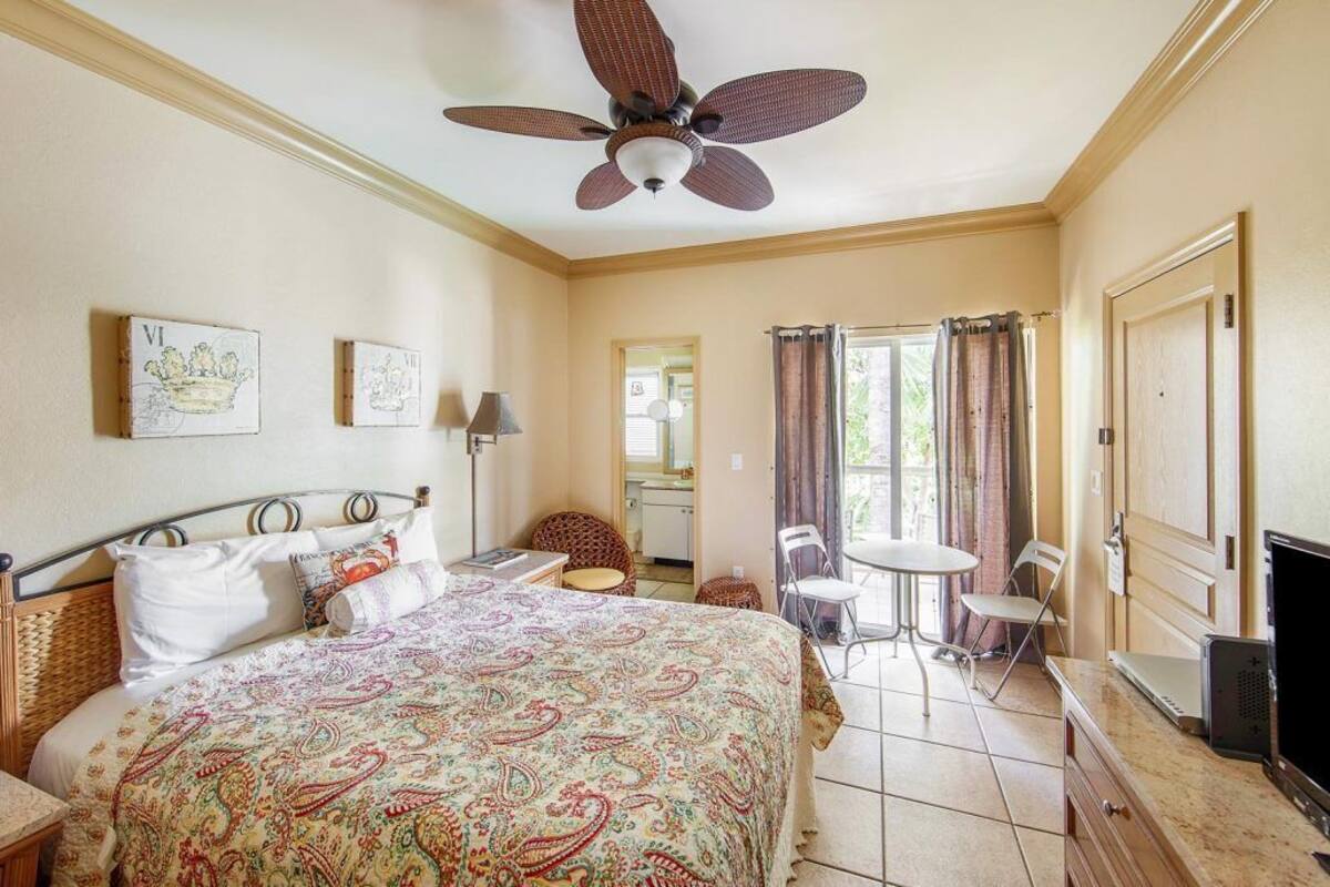 A hotel room with a bed, a ceiling fan, a small table, two chairs, a TV, and a window with curtains.