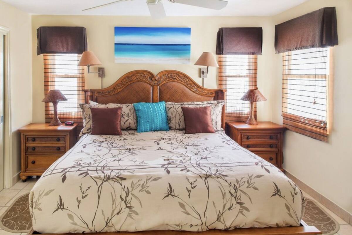 A bedroom with a large bed, two bedside tables, a ceiling fan, and a painting of a beach.