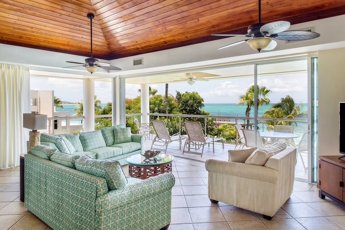 A spacious living area with a large sofa, a smaller sofa, and a coffee table. There are ceiling fans, a TV, and a view of the ocean.