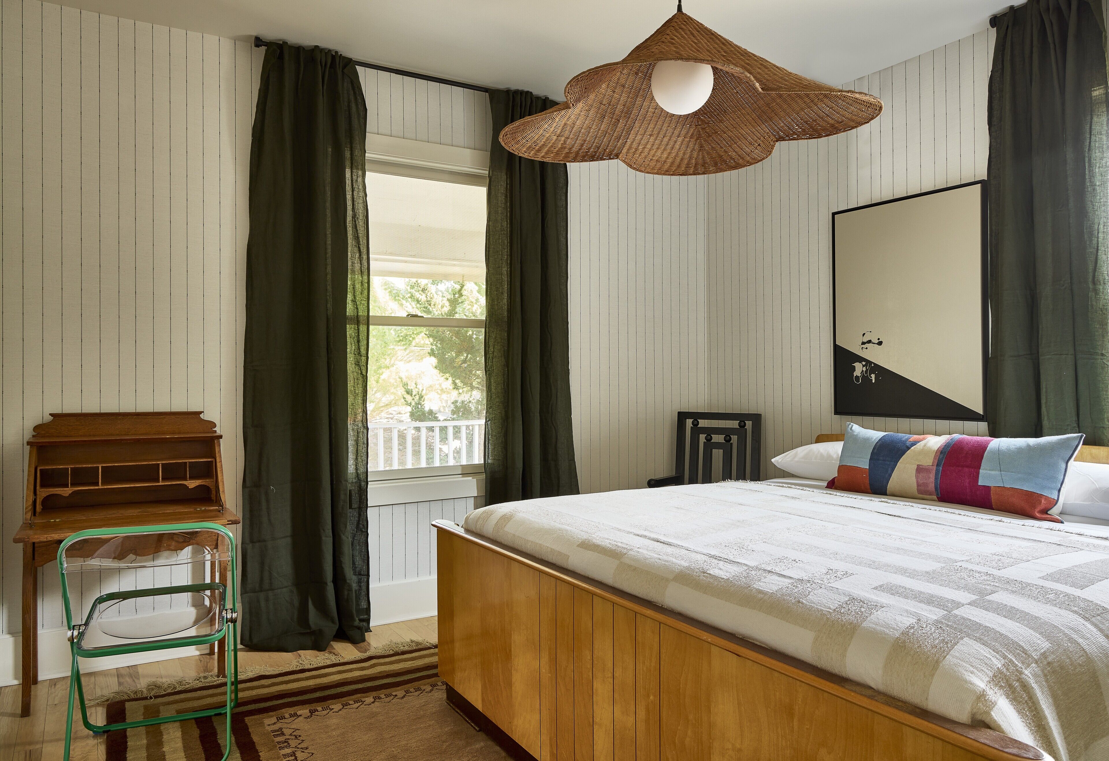 A bedroom with a wooden bed, a green metal chair, a wicker pendant light, a large mirror, and a window with curtains.