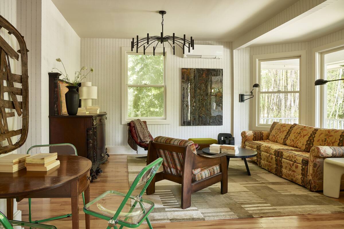 Un salon chaleureux avec une table basse en bois, un canapé et un fauteuil. On y trouve une bibliothèque, une lampe et une grande fenêtre qui laisse entrer la lumière naturelle.