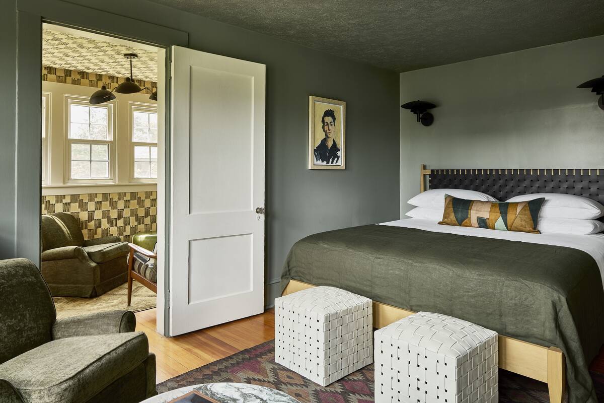 Une chambre à coucher avec un lit, deux poufs, une chaise, une fenêtre et un tableau au mur.