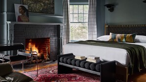 A bedroom with a fireplace, a bed with a green bedspread, a red patterned rug, and a large window with curtains. - Blind Tiger Asheville (Asheville)