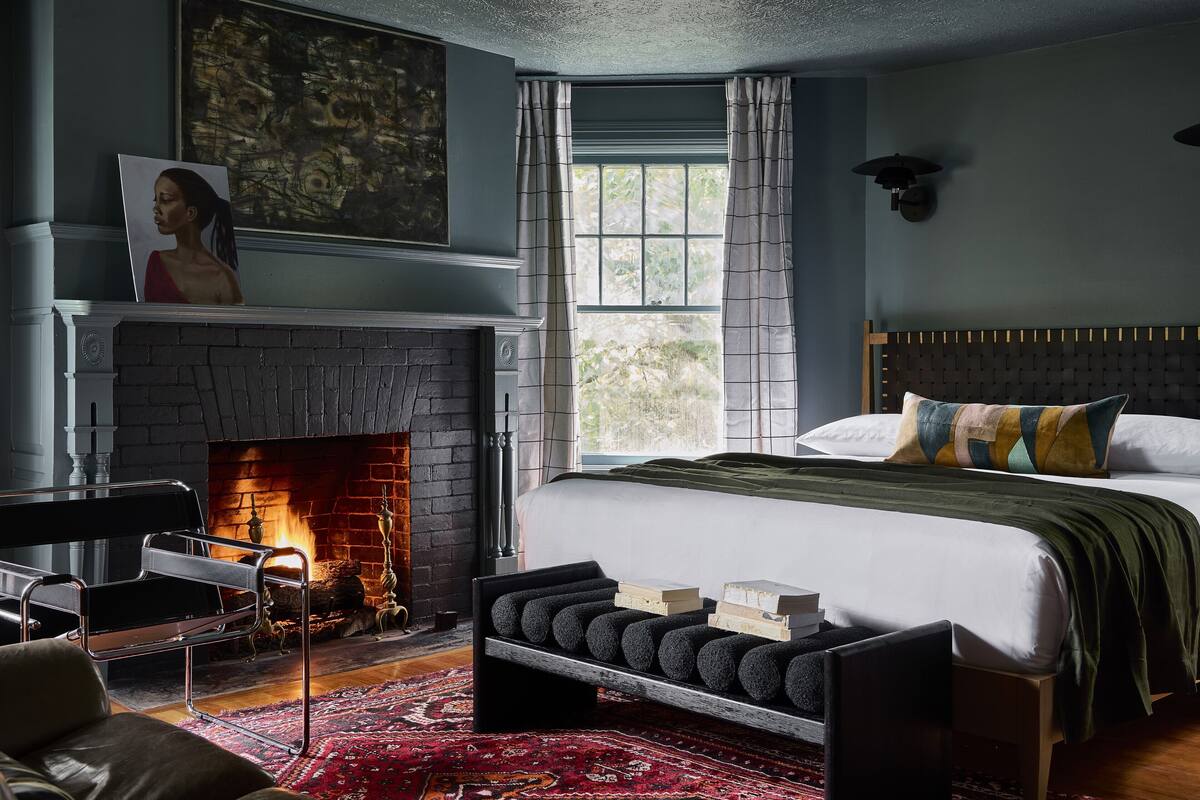 Une chambre avec une cheminée, un lit recouvert d’une courtepointe verte, un tapis rouge à motifs et une grande fenêtre avec des rideaux.
