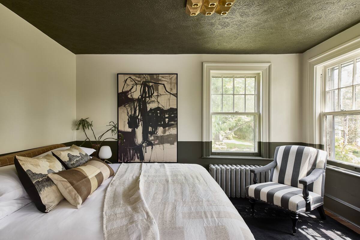 Une chambre avec un grand lit, un radiateur, un fauteuil à rayures et une fenêtre donnant sur un paysage verdoyant.