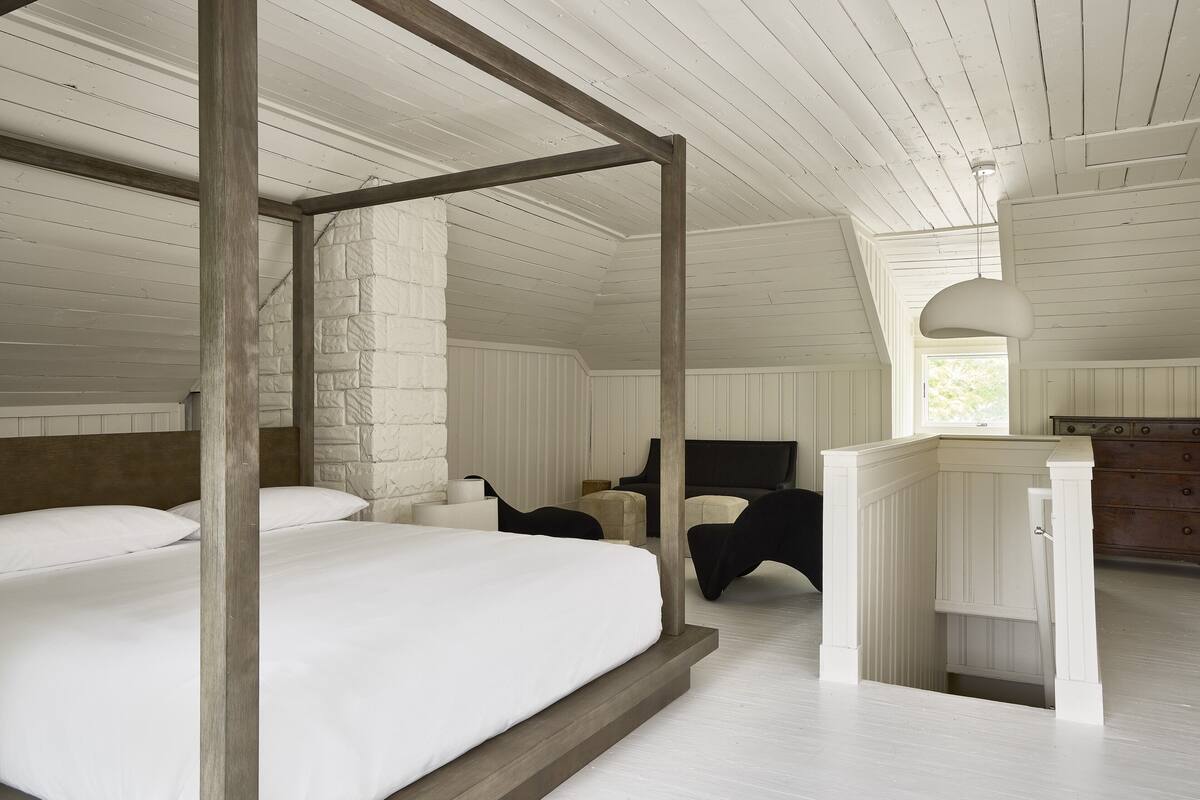 Une chambre à coucher avec un lit à baldaquin, une tête de lit en bois, un mur d’accent en briques blanches, une commode en bois et une grande fenêtre.