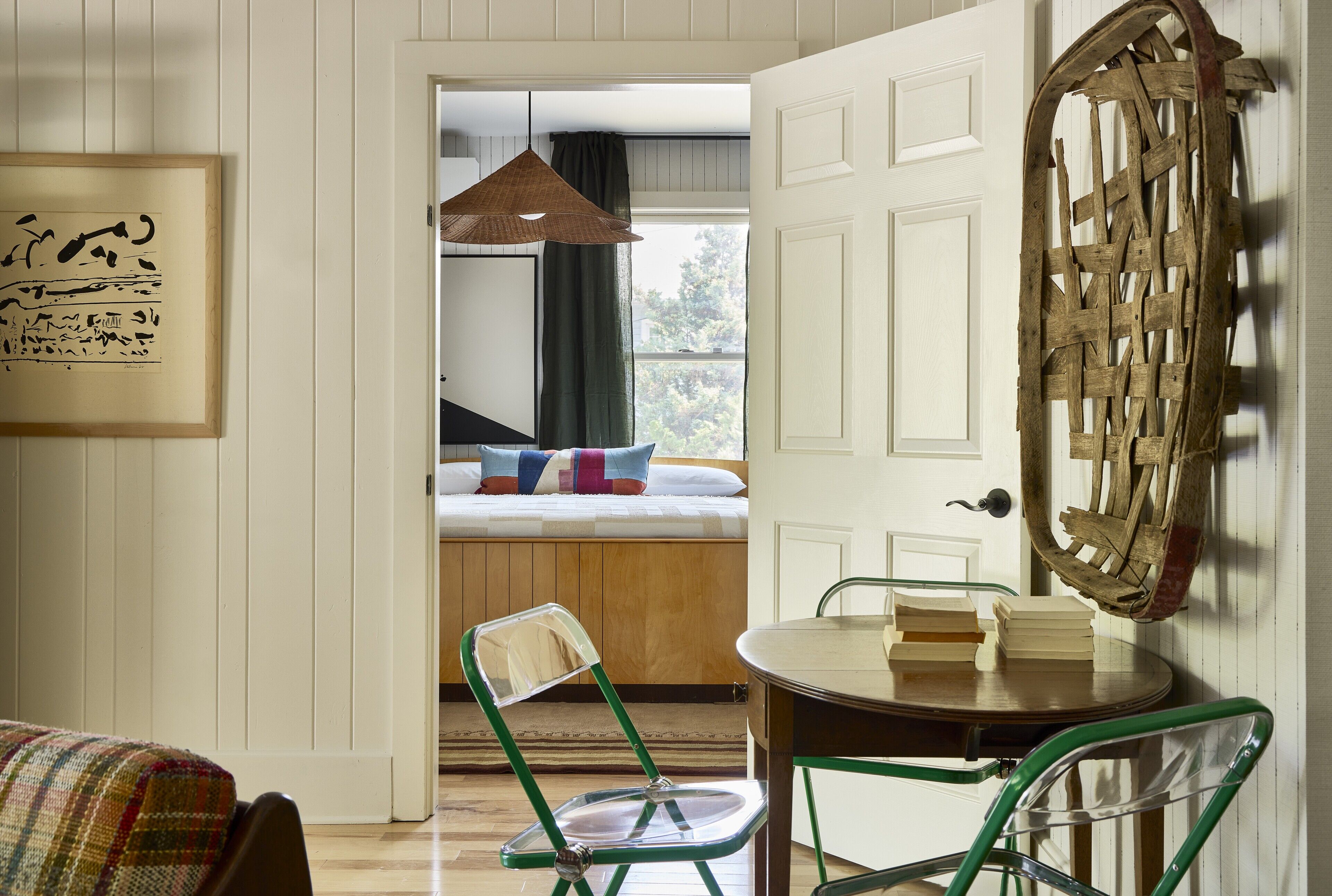 A room with a bed, a chair, a table with books, a hanging light, and a door with a woven decoration.