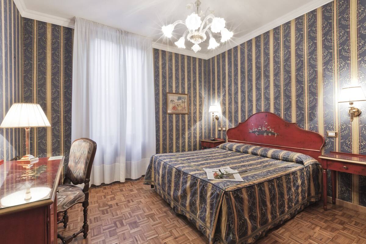Een hotelkamer met een houten vloer, een bed met een gestreept dekbedovertrek, een bureau met een lamp, een stoel en een nachtkastje met een lamp.