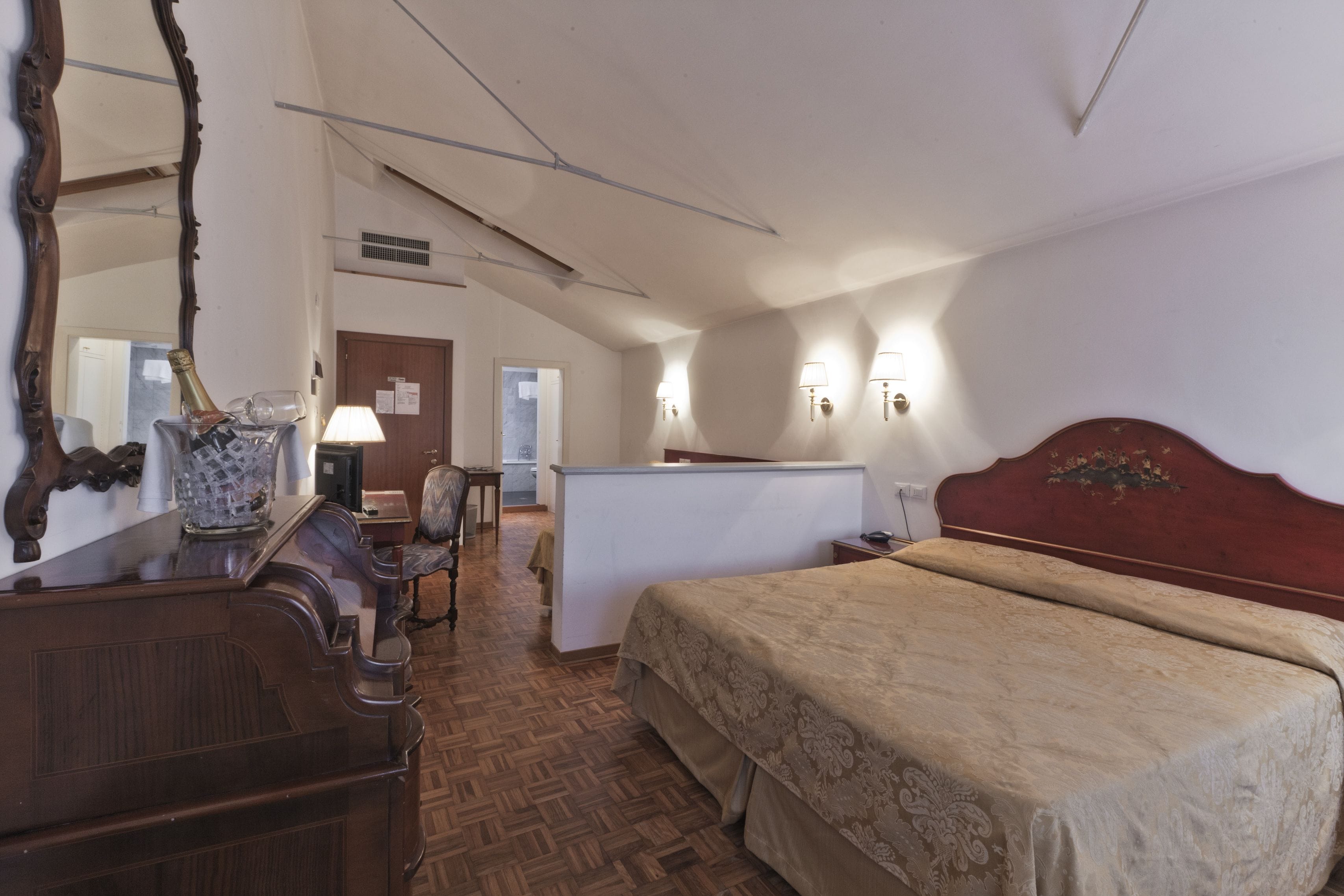 Ein Hotelzimmer mit einem Holzkoffer, einem Bett mit Kopfteil, einem Schreibtisch mit Stuhl und einer kleinen Küchenzeile.