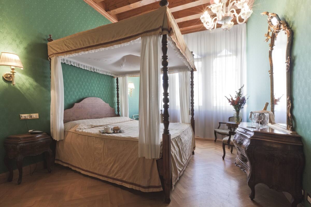 Een hemelbed met houten frame en witte beddengoed, een nachtkastje van hout met een lamp, een dressoir met spiegel en een kroonluchter.