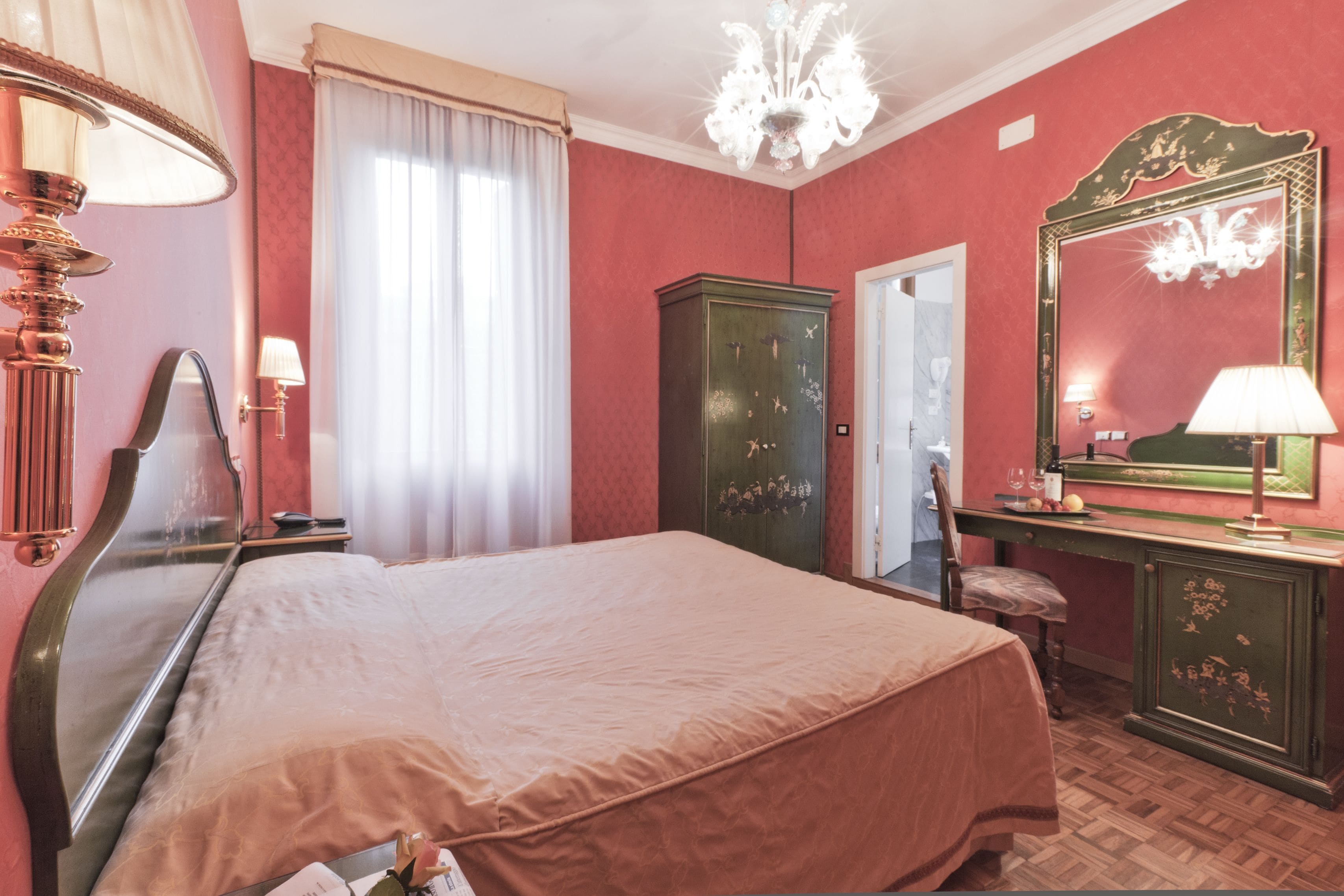 Une chambre avec un lit, une commode, un miroir, un lustre et une fenêtre avec des rideaux.