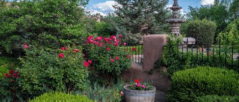 Un jardín con una fuente de piedra, flores rojas y diversos arbustos.