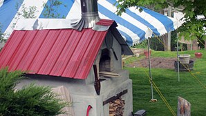A small outdoor structure with a metal roof, a chimney, and a stack of wood. - Inn at Neshobe River (Brandon)