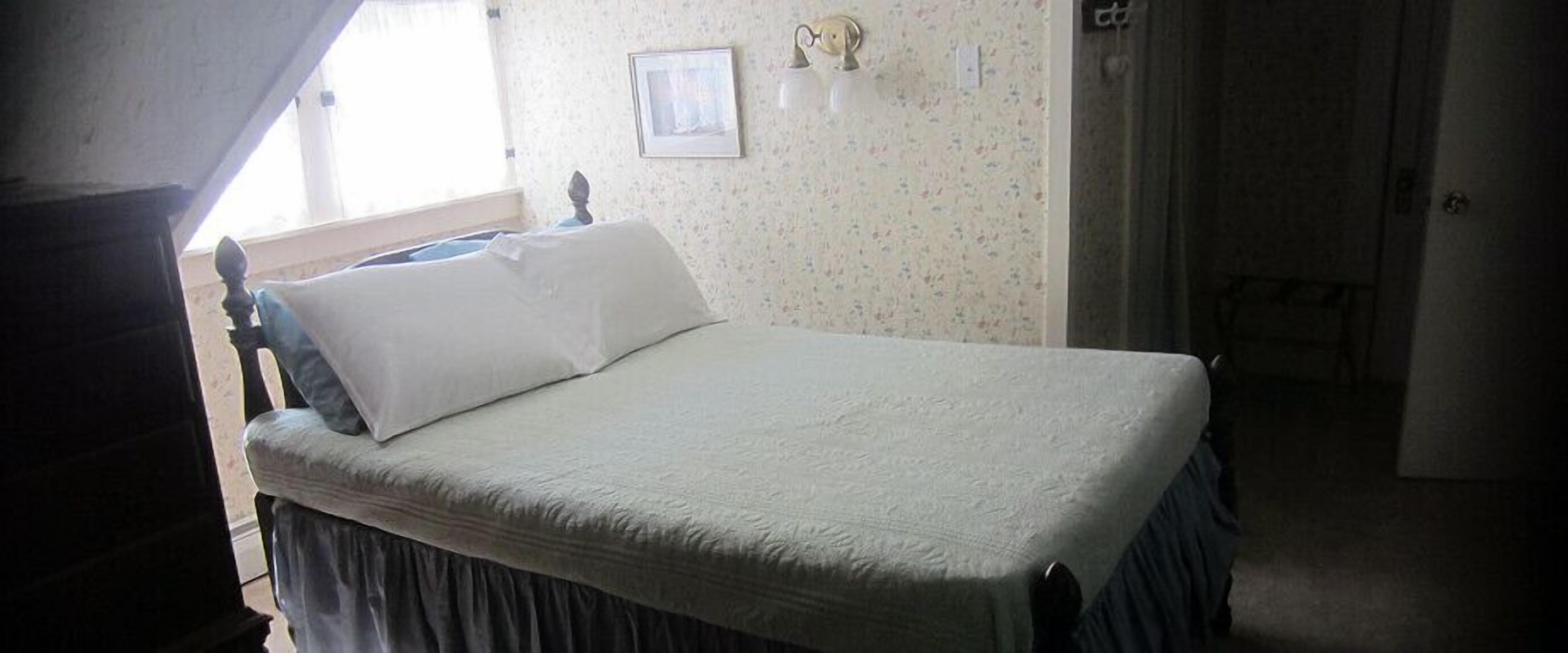 A bed with a white bedspread and a dark skirted headboard in a room with floral wallpaper and a framed picture on the wall.