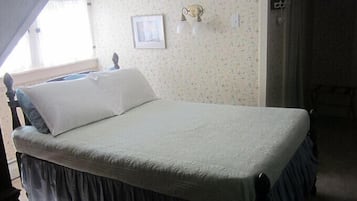 Un lit recouvert d’une courtepointe blanche, avec une tête de lit sombre et ornée d’une jupe, dans une pièce dont les murs sont recouverts d’un papier peint à motifs floraux et où est accroché un tableau encadré.
