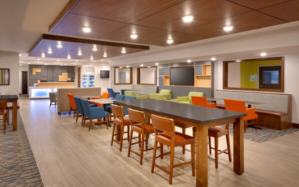 A modern hotel lobby with a long dining table, multiple seating areas, and a reception desk.