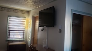 A room with a television mounted on the wall, a window with blinds, a wooden door, and a small table with a lamp. - Northside Motel (Williamstown)