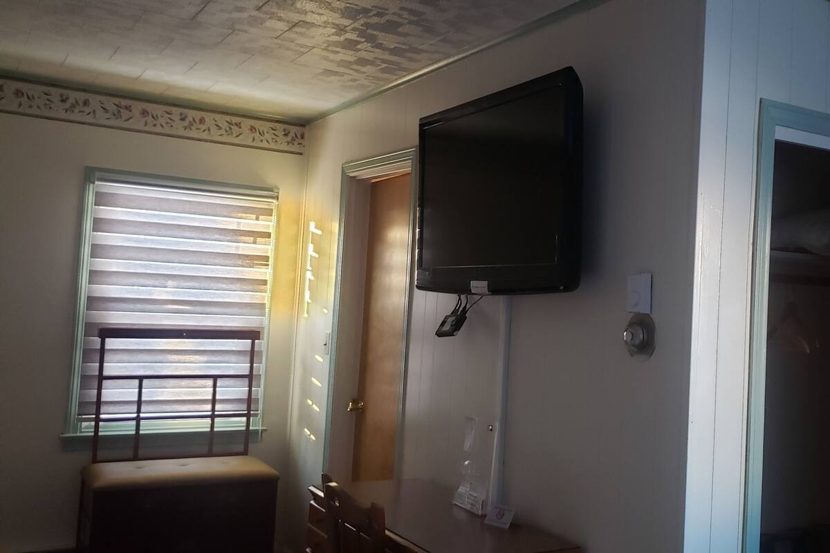 A room with a television mounted on the wall, a window with blinds, a wooden door, and a small table with a lamp.