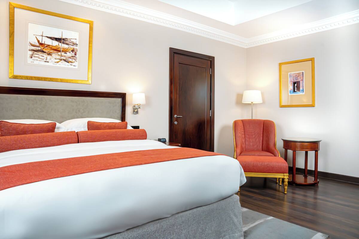 A hotel room with a large bed, a chair, a door, and two framed pictures on the walls.