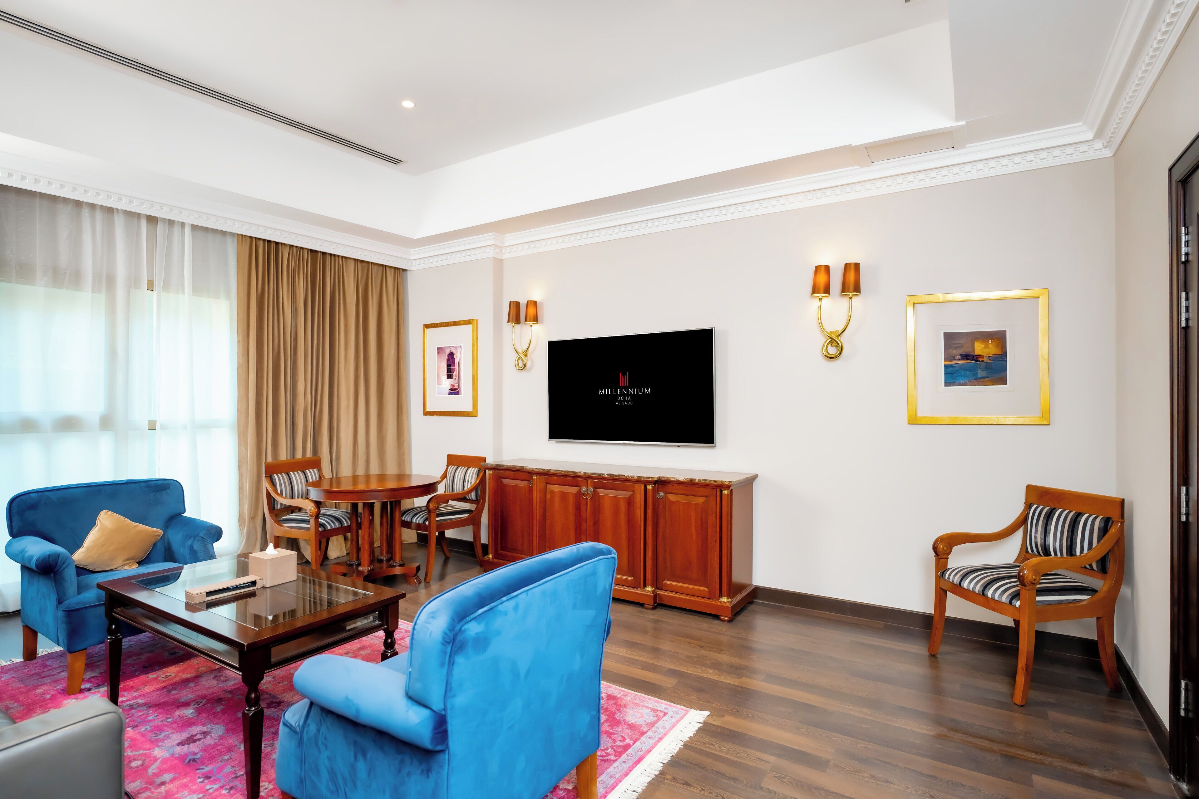 Una sala de estar moderna con un televisor de pantalla plana grande, sillones azules, una mesa de centro de madera y una alfombra roja estampada.