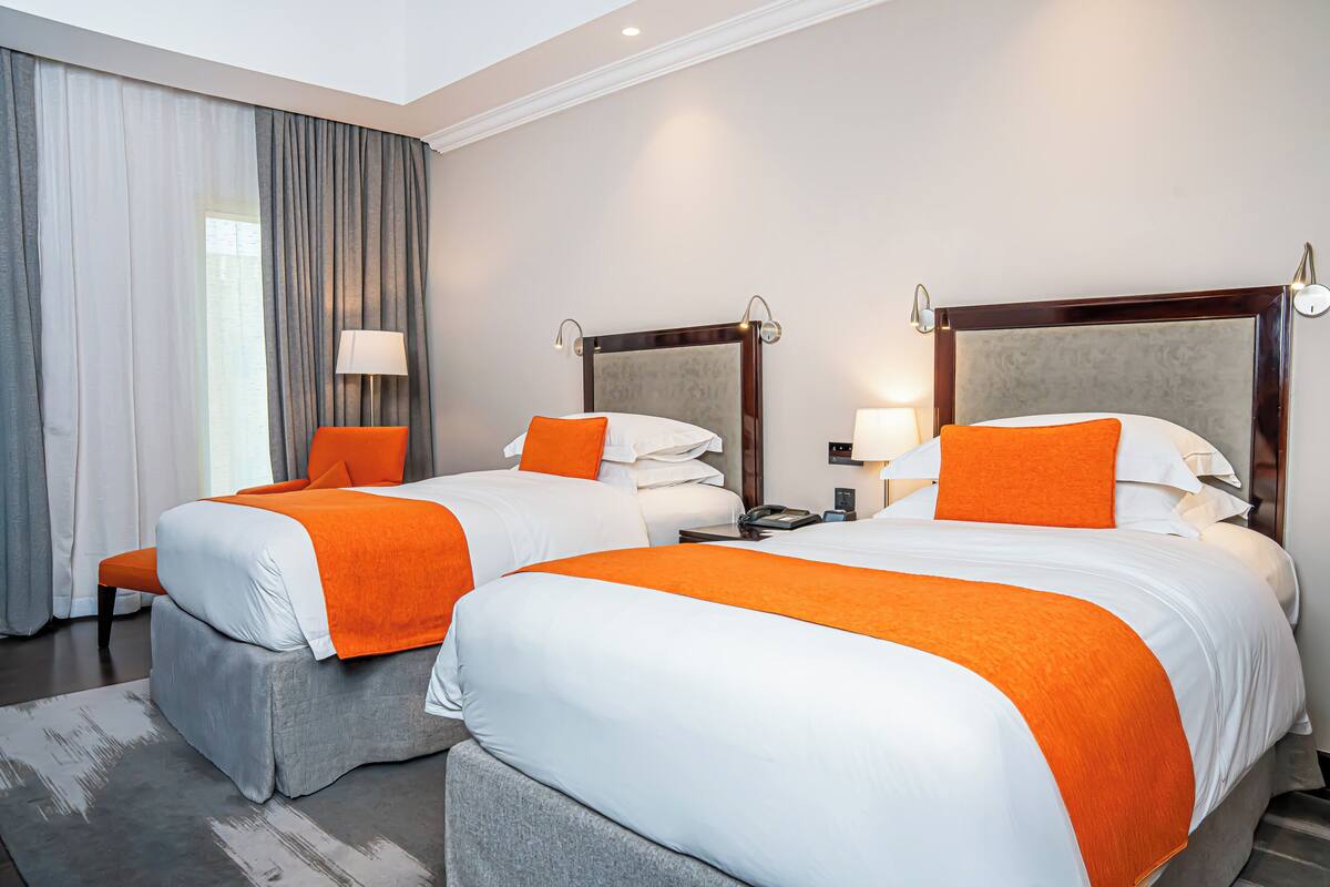 A hotel room with two beds, each with white and orange bedding, a grey upholstered bench, a grey rug, and a large window with grey curtains.