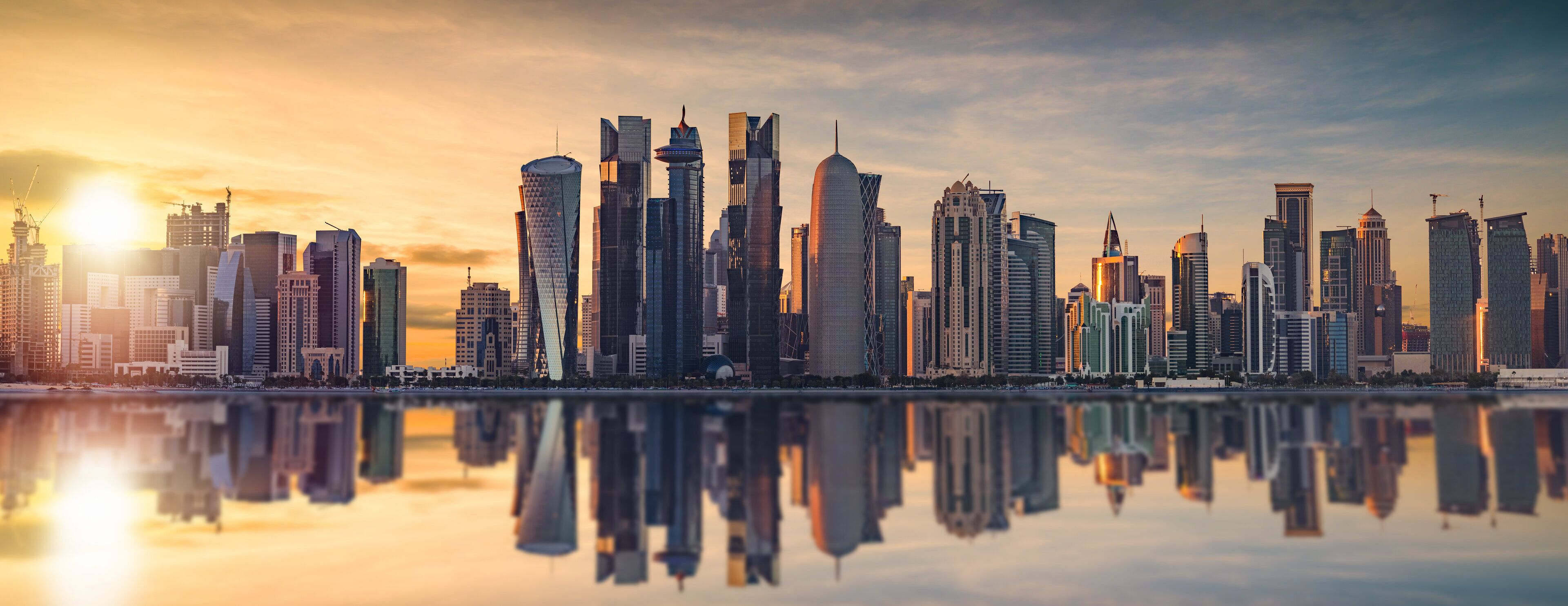 A city skyline with modern high-rise buildings reflected in a calm body of water during sunset.