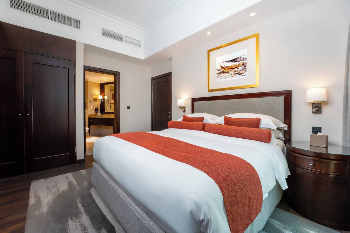 A hotel room with a large bed, a nightstand, a framed picture on the wall, and a door leading to another room.