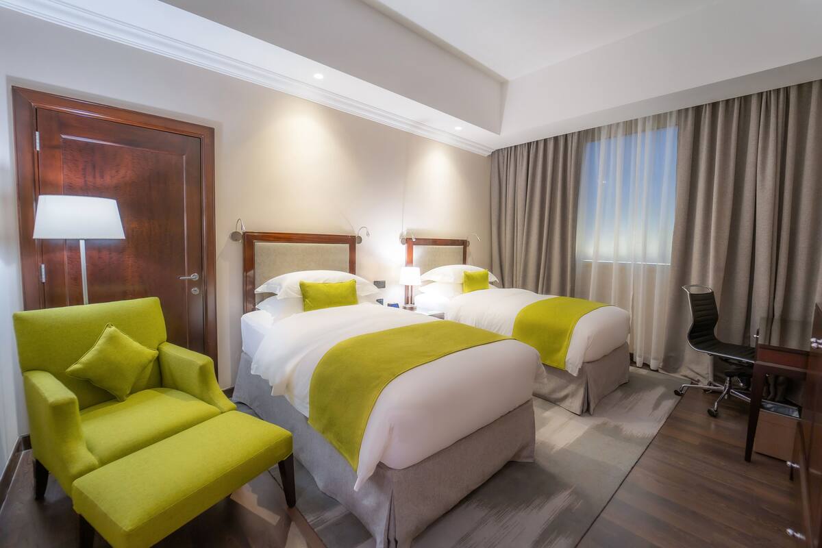 A hotel room with two beds, a green armchair, a desk, and a large window with curtains.