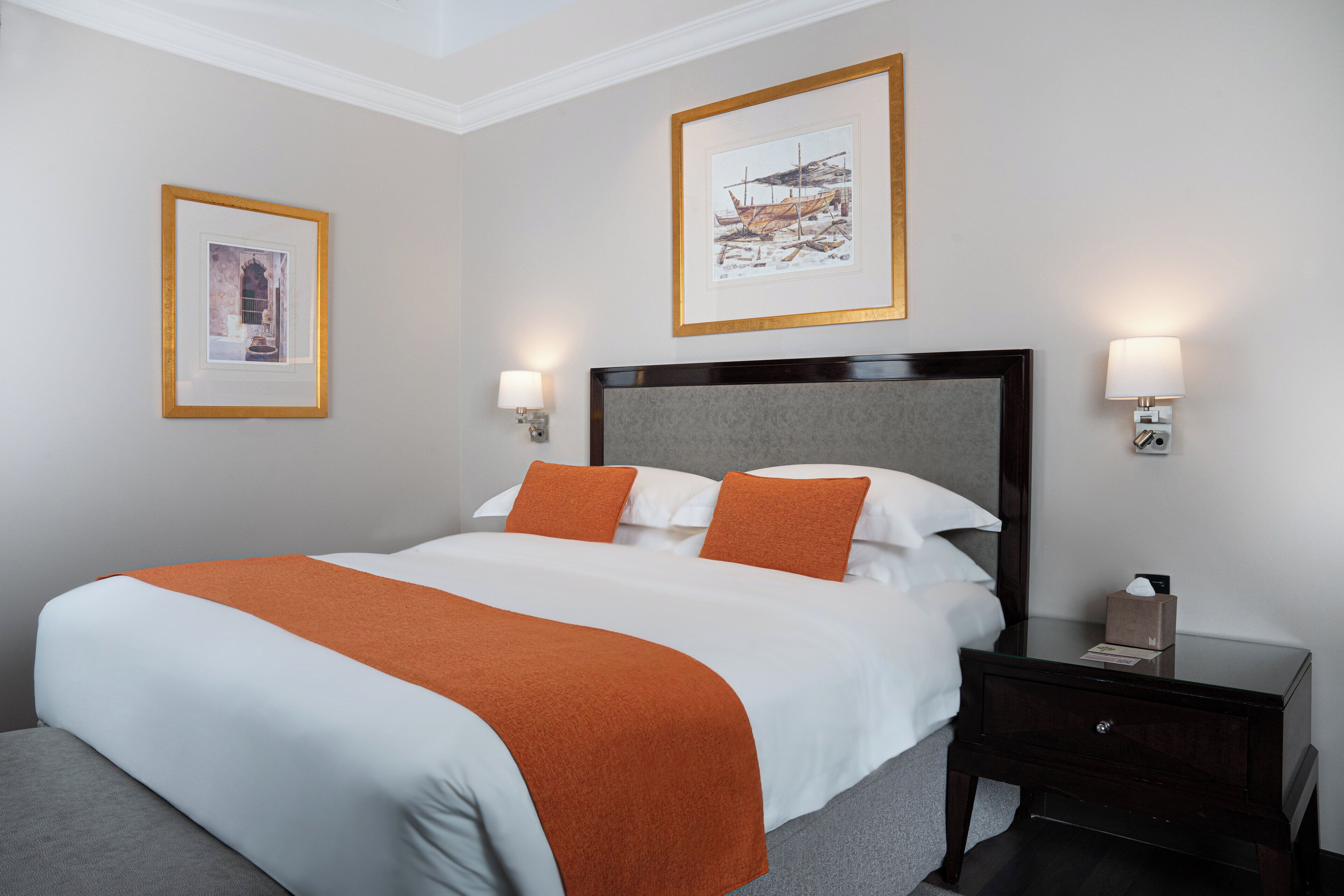 Una cama bien hecha con ropa de cama blanca y naranja, un cabecero de madera oscura y dos lámparas fijadas a la pared.