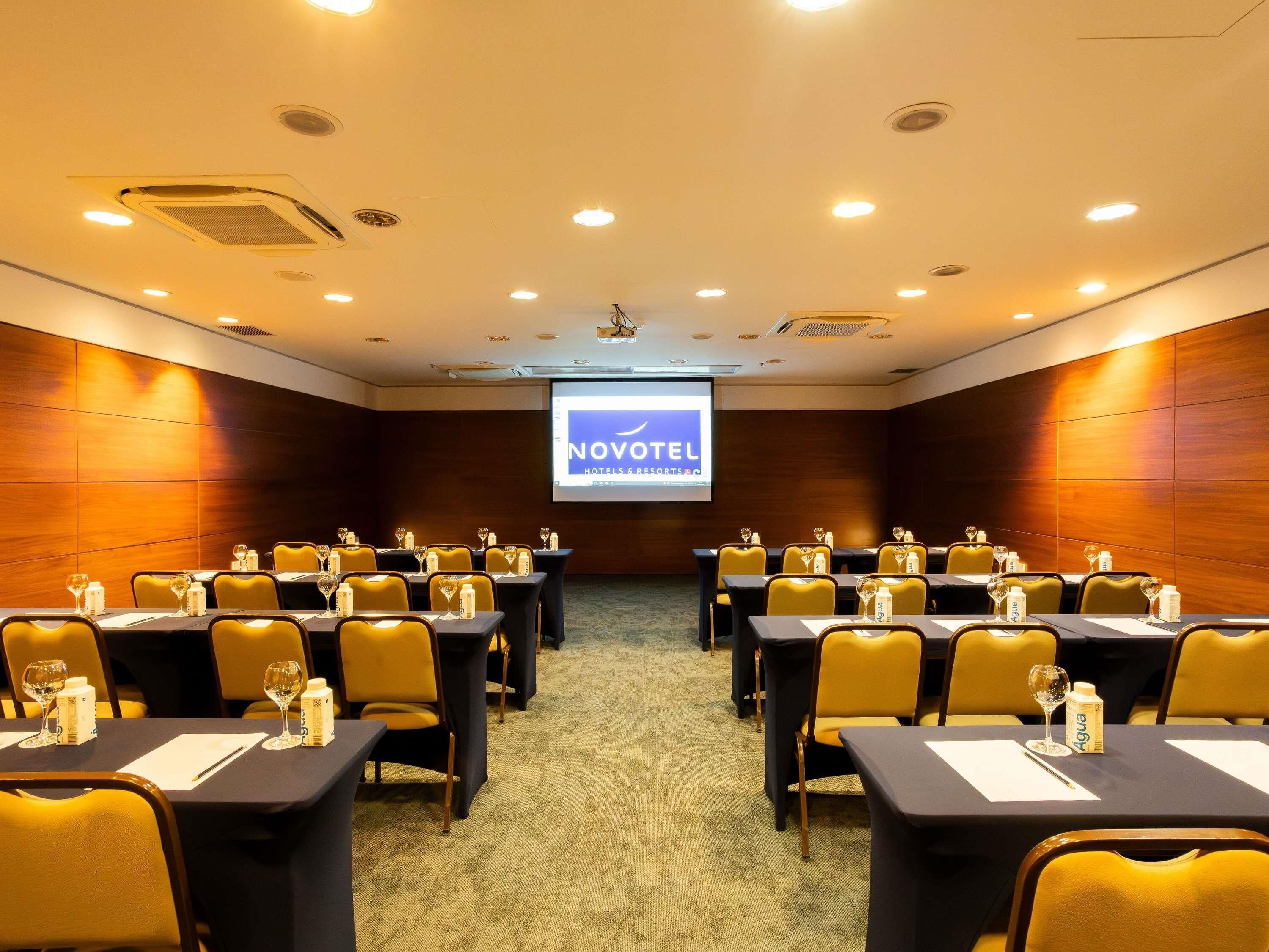 Un salón de conferencias con mesas, sillas y una pantalla de proyector que muestra 'NOVOTEL Hotels & Resorts'.
