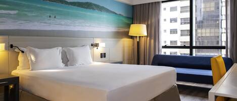 A hotel room with a large bed, a desk, a chair, a lamp, and a view of a beach through a window.