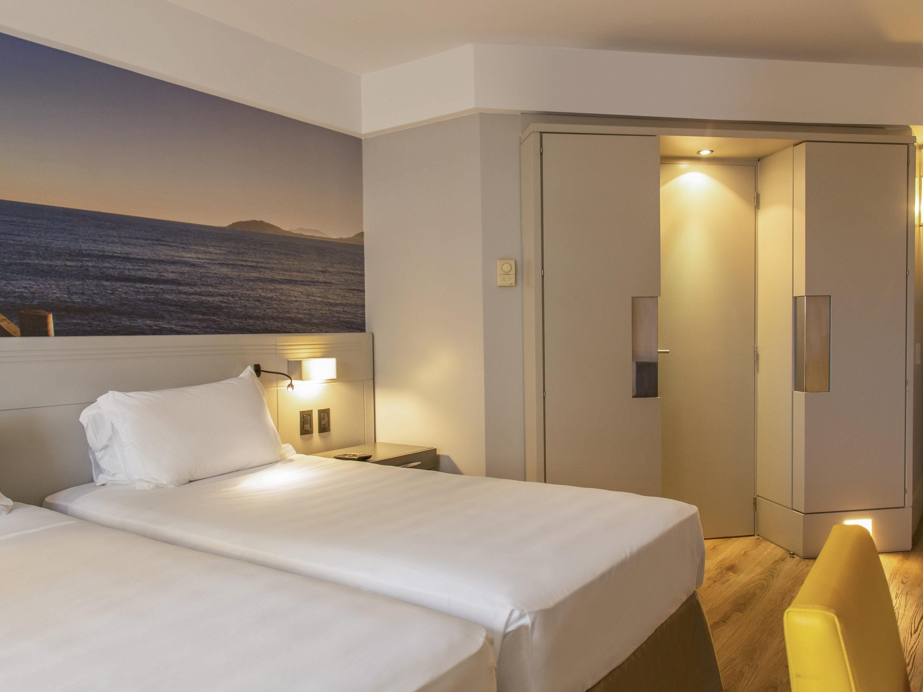 A hotel room with two beds, a nightstand, a lamp, a wardrobe, and a painting of a landscape.