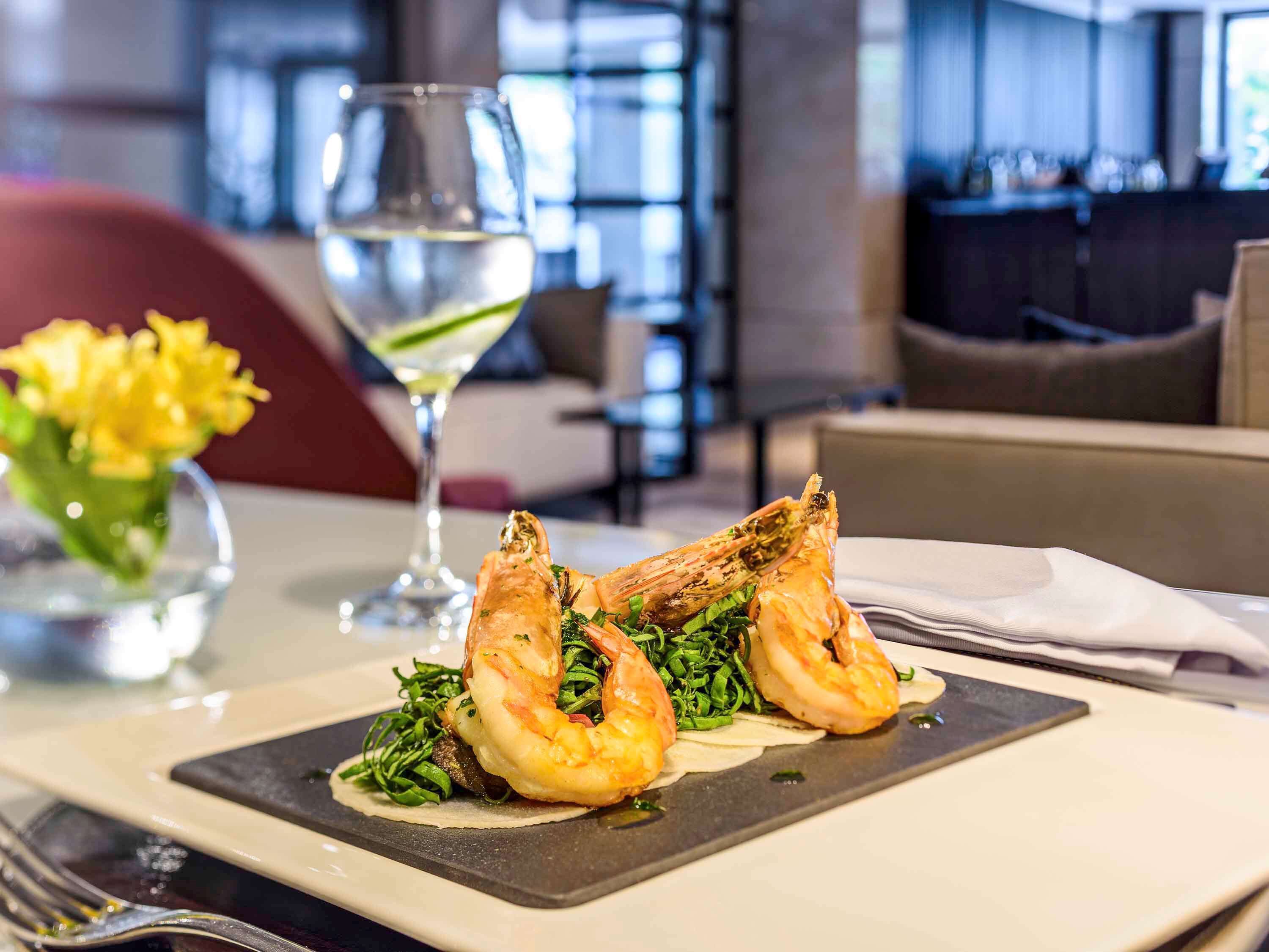 A dining table with a plate of grilled shrimp and greens, a glass of white wine, and a vase of yellow flowers.
