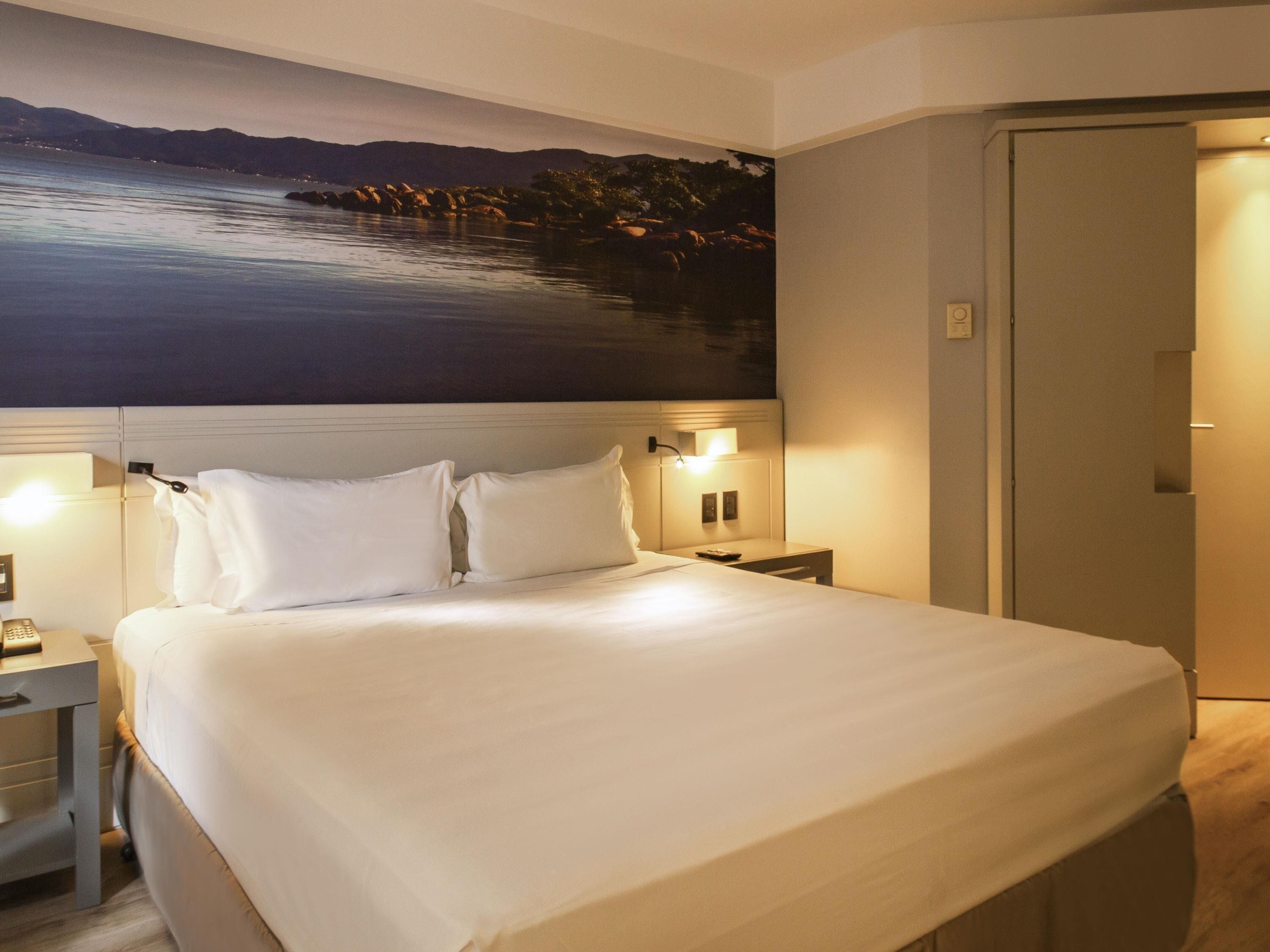 A hotel room with a large bed, a nightstand with a lamp, a wall-mounted TV, and a door.