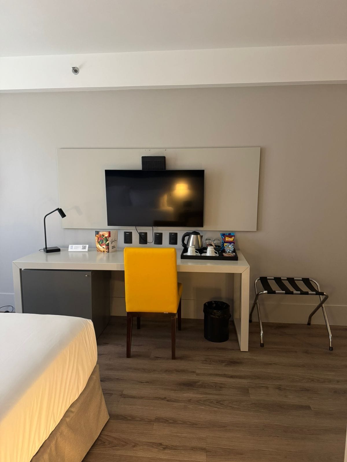 Habitación de hotel con cama, escritorio con televisor, silla, kitchenette pequeña y un basurero.