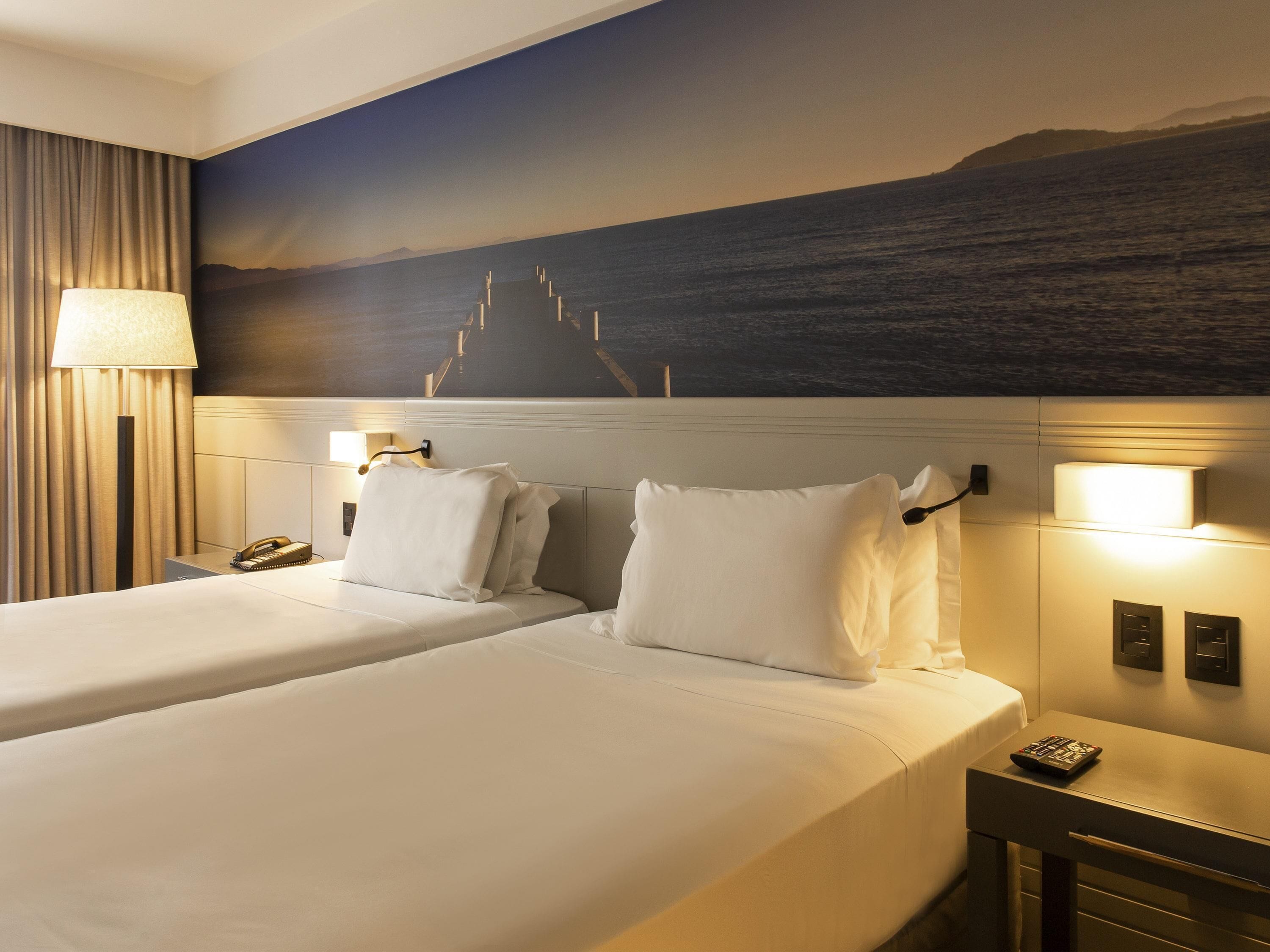 A hotel room with two beds, a nightstand, a lamp, and a wall mural.