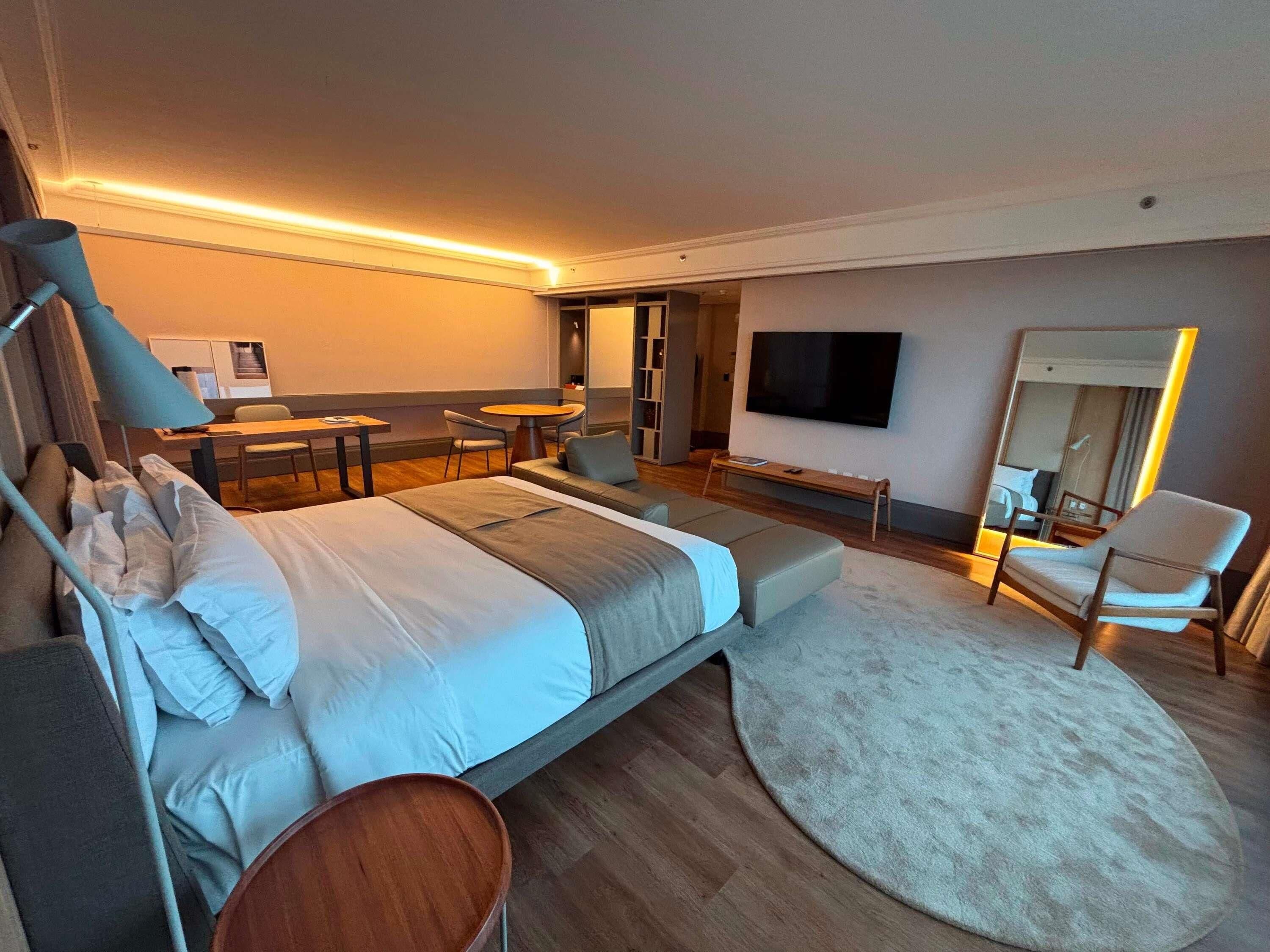 Habitación de hotel moderna con cama, escritorio, silla, televisor y un espejo grande.