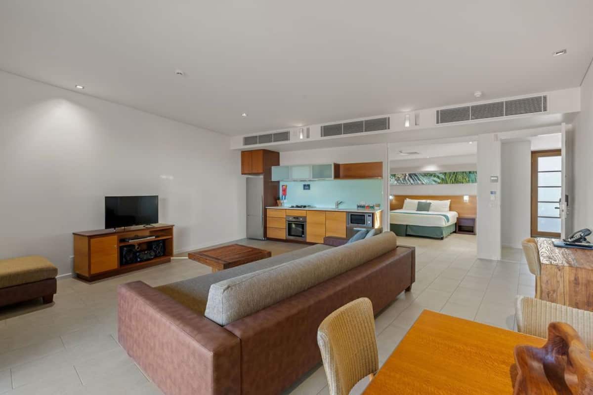 Een moderne woonkamer met een bank, televisie en een eethoek. In de achtergrond is een keuken zichtbaar, met een bed en een balkon.