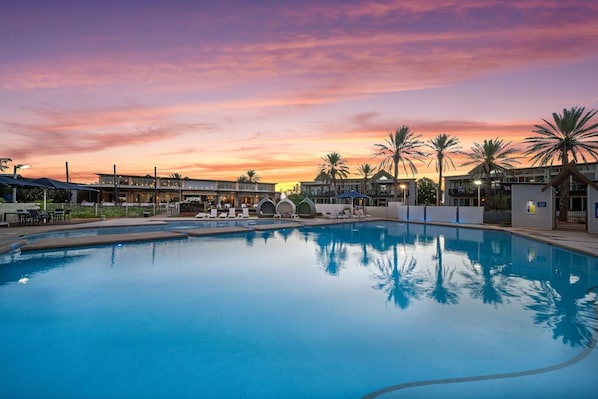 A swimming pool with lounge chairs and palm trees, surrounded by buildings with a sunset sky in the background. - Mantarays Ningaloo Beach Resort (Exmouth)