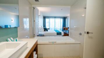 A modern hotel bathroom with a large bathtub, a separate shower area, and a visible bedroom with a bed and curtains.