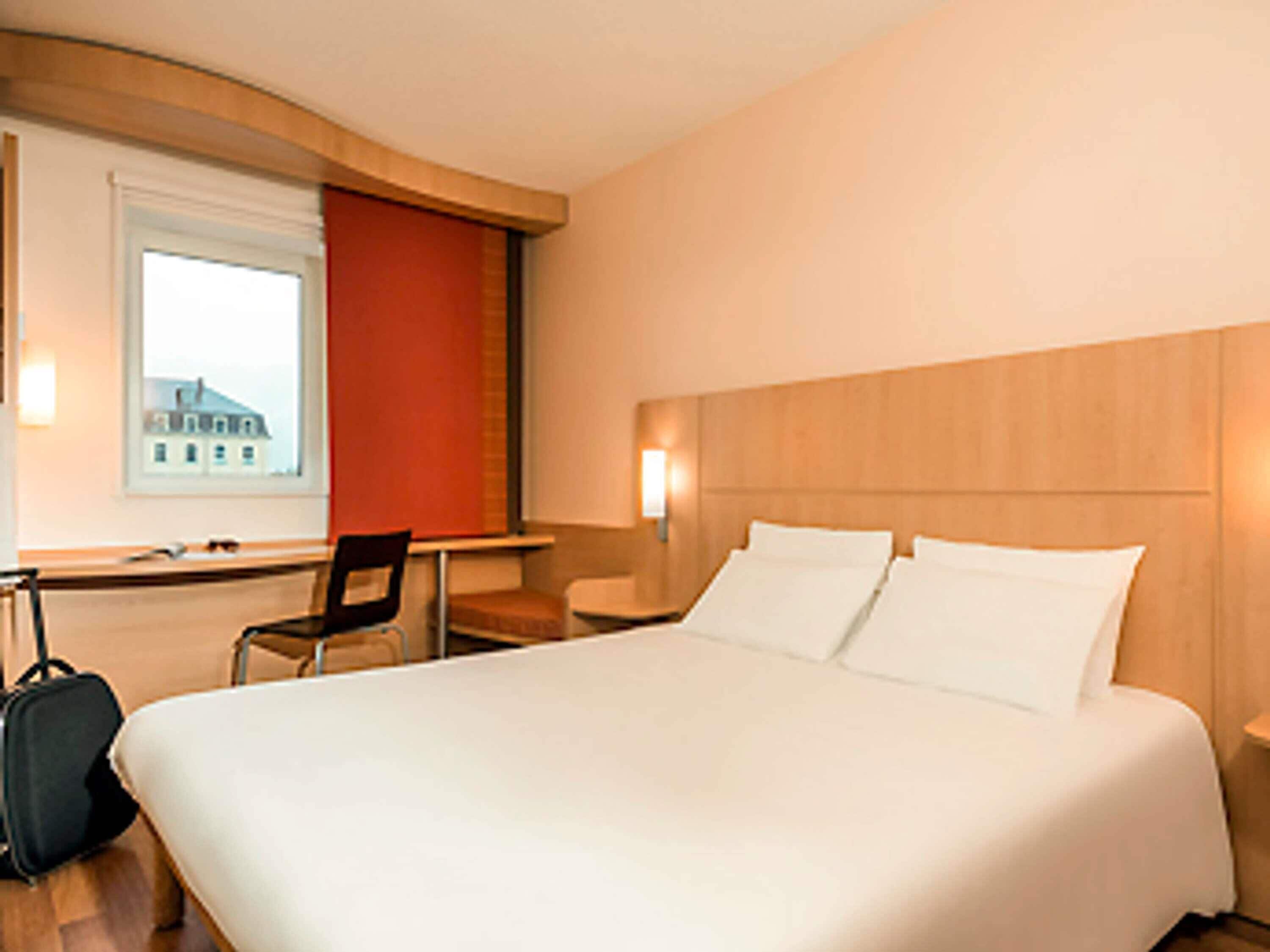 Quarto de hotel com uma cama grande, uma escrivaninha, uma cadeira e uma mala.
