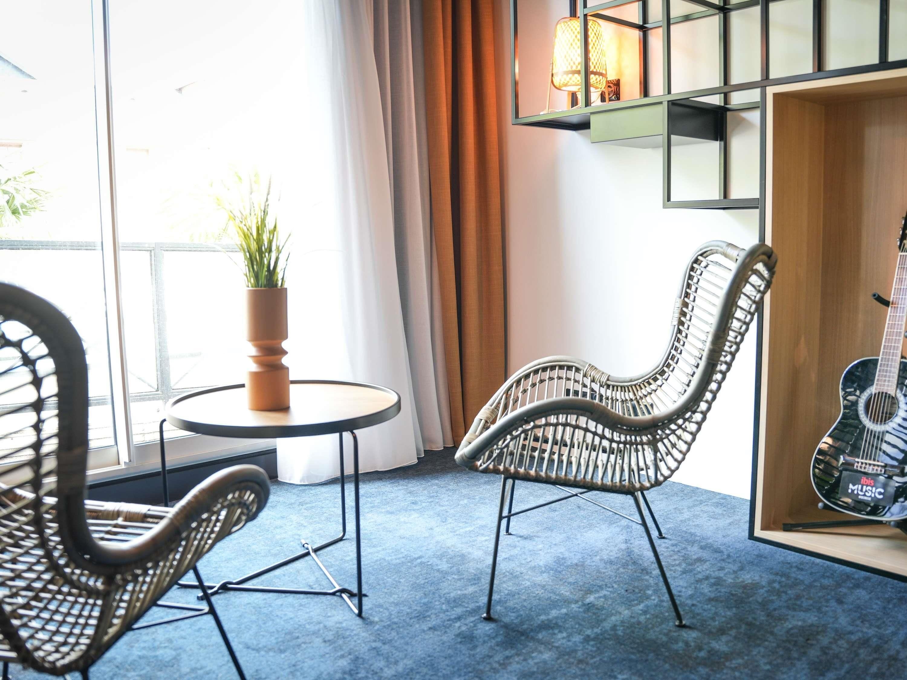 A room with wicker chairs, a small round table, a potted plant, a guitar, and a wall-mounted shelf with a lamp.