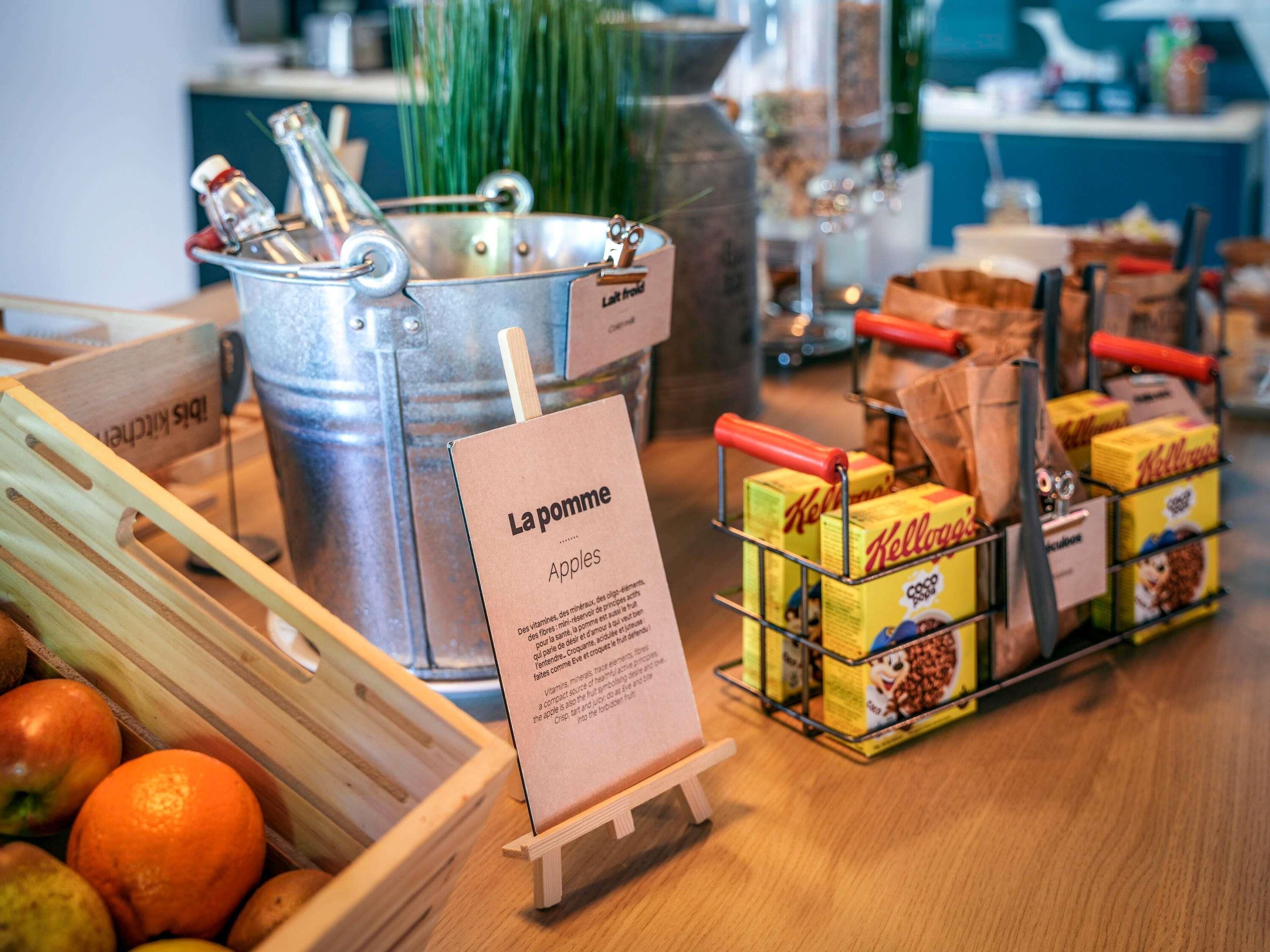 Eine Küchenarbeitsplatte mit einem Metallkübel mit dem Aufdruck 'La pomme', einer Holzkiste voller Äpfel und einer Auswahl an Kellogg's-Müsli-Kartons.