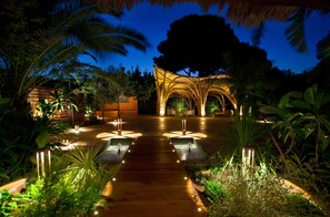 A well-lit pathway leading to a covered area with arches, surrounded by lush greenery and palm trees. - Hotel Les Rotes (Denia)