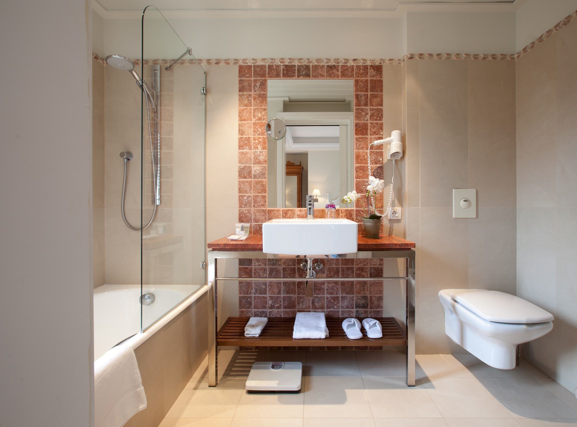 A modern bathroom with a glass-enclosed shower, a white sink, a toilet, and a bench with towels and slippers.