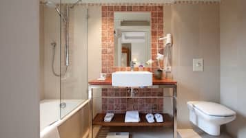 Un baño moderno con ducha de vidrio, lavamanos blanco, inodoro y banco con toallas y zapatillas.