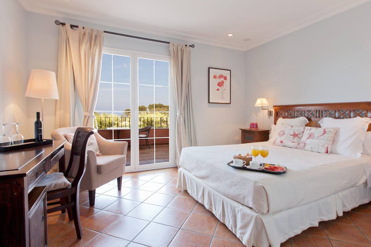 A hotel room with a bed, a desk, a chair, a nightstand, and a view through a glass door.