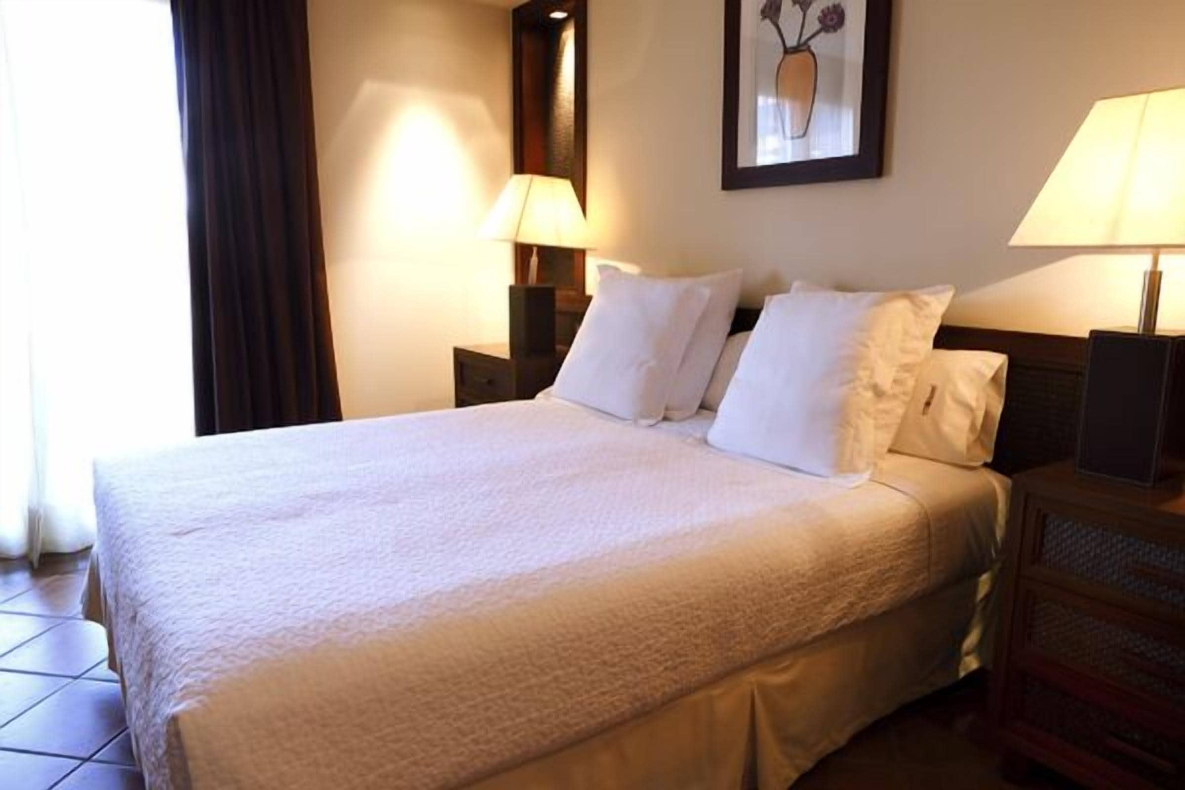 A neatly made bed with white linens, two bedside tables with lamps, a framed picture on the wall, and a window with curtains.