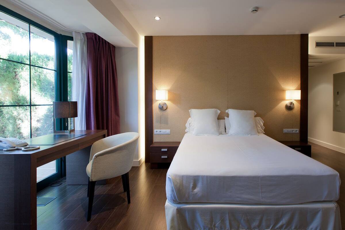 A hotel room with a large bed, a desk with a chair, a bedside table, and a window with curtains.