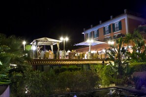 A nighttime outdoor area with a covered seating area, a pool, and a building with multiple windows. - Hotel Les Rotes (Denia)